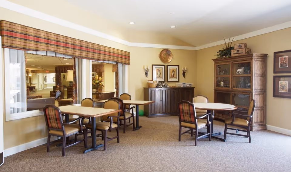 A cozy interior room with beige walls and carpeted floor featuring two tables surrounded by chairs with plaid upholstery. There is a wooden cabinet with glass doors on the right side, decorative wall art, and a sideboard with framed pictures and wall sconces above it. Large windows with plaid valances look into another room.