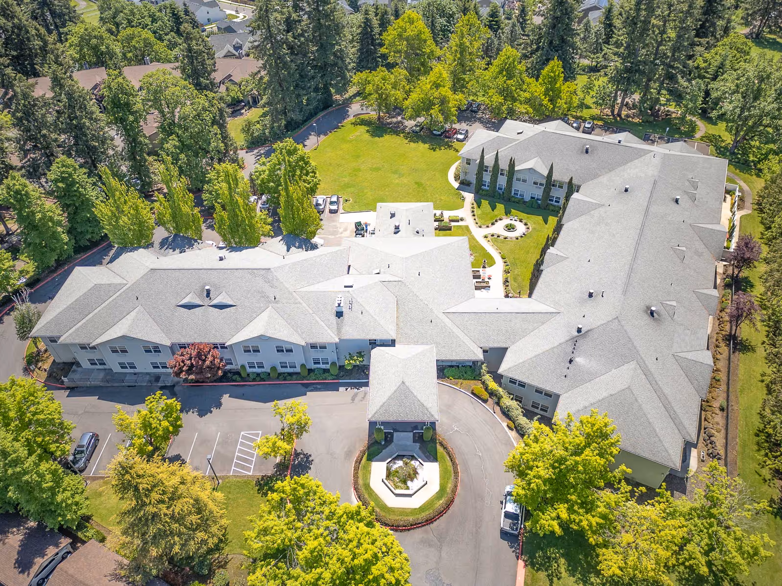 Aerial view of Tanner Spring Assisted Living & Memory Care facility showing a large building complex surrounded by green trees and lawns, with a circular driveway and parking spaces visible.