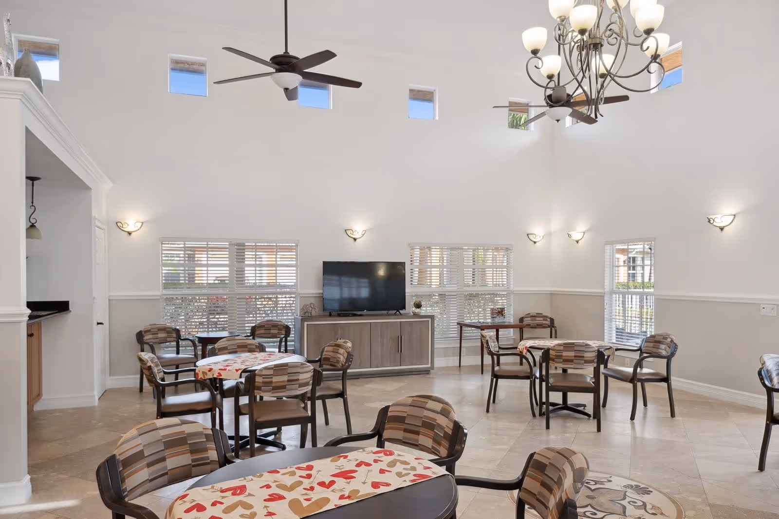 Bright, spacious common dining/lounge area with round tables and patterned chairs, a TV console, high ceilings with fans and chandelier, and large windows.