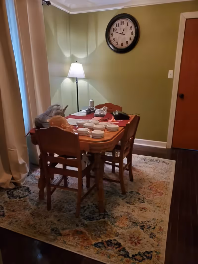 A dining room with a wooden table and four chairs on a patterned rug. The table has several round white containers, a lamp on a side table, a large wall clock showing 9:15, and a closed wooden door. The walls are painted green and there are beige curtains on the left side.