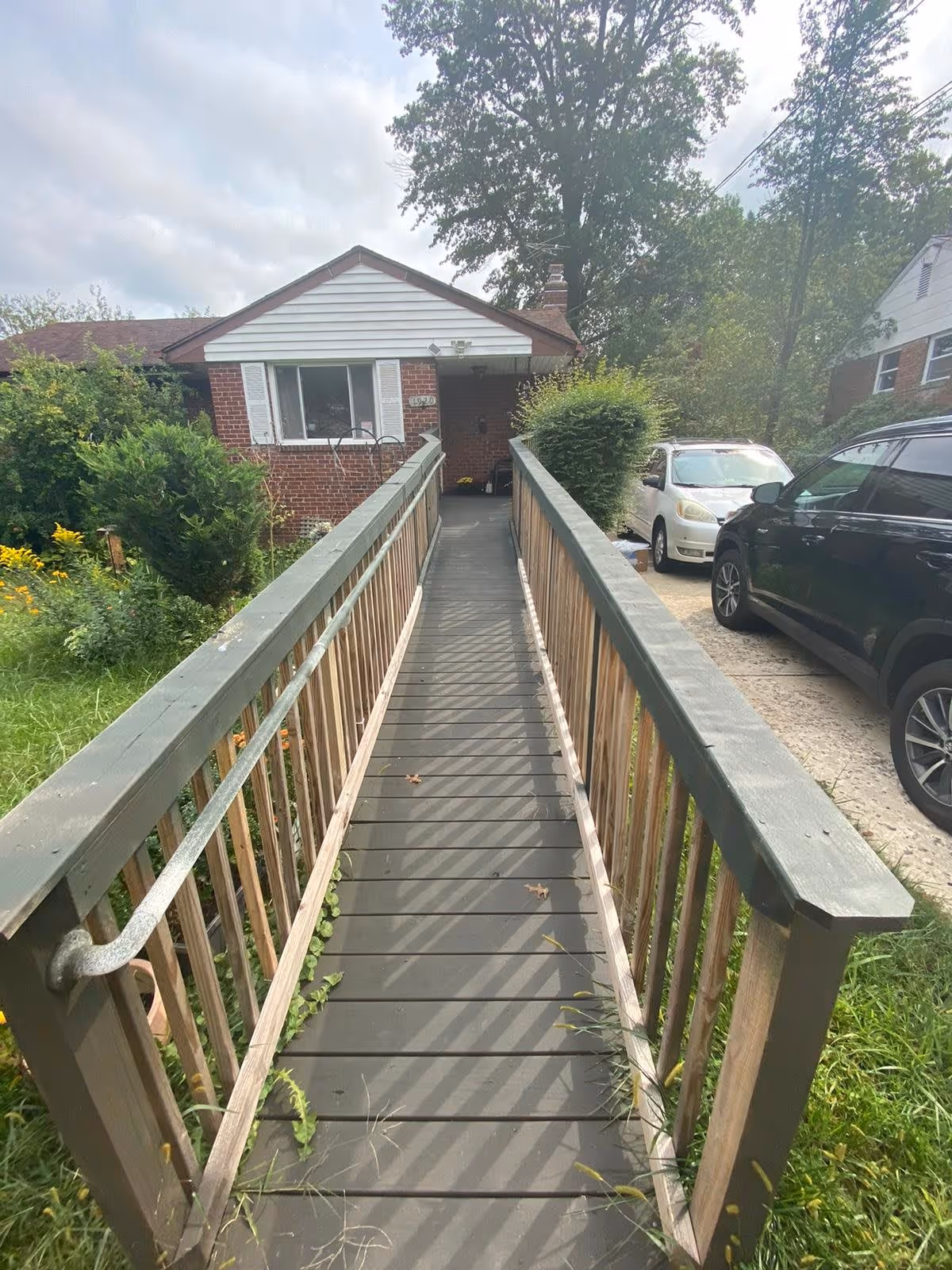 A wooden wheelchair ramp with handrails on both sides leading up to the front entrance of a single-story brick house with white siding and a window with white shutters. There are bushes and greenery on either side of the ramp, and two cars parked on a driveway to the right of the house.