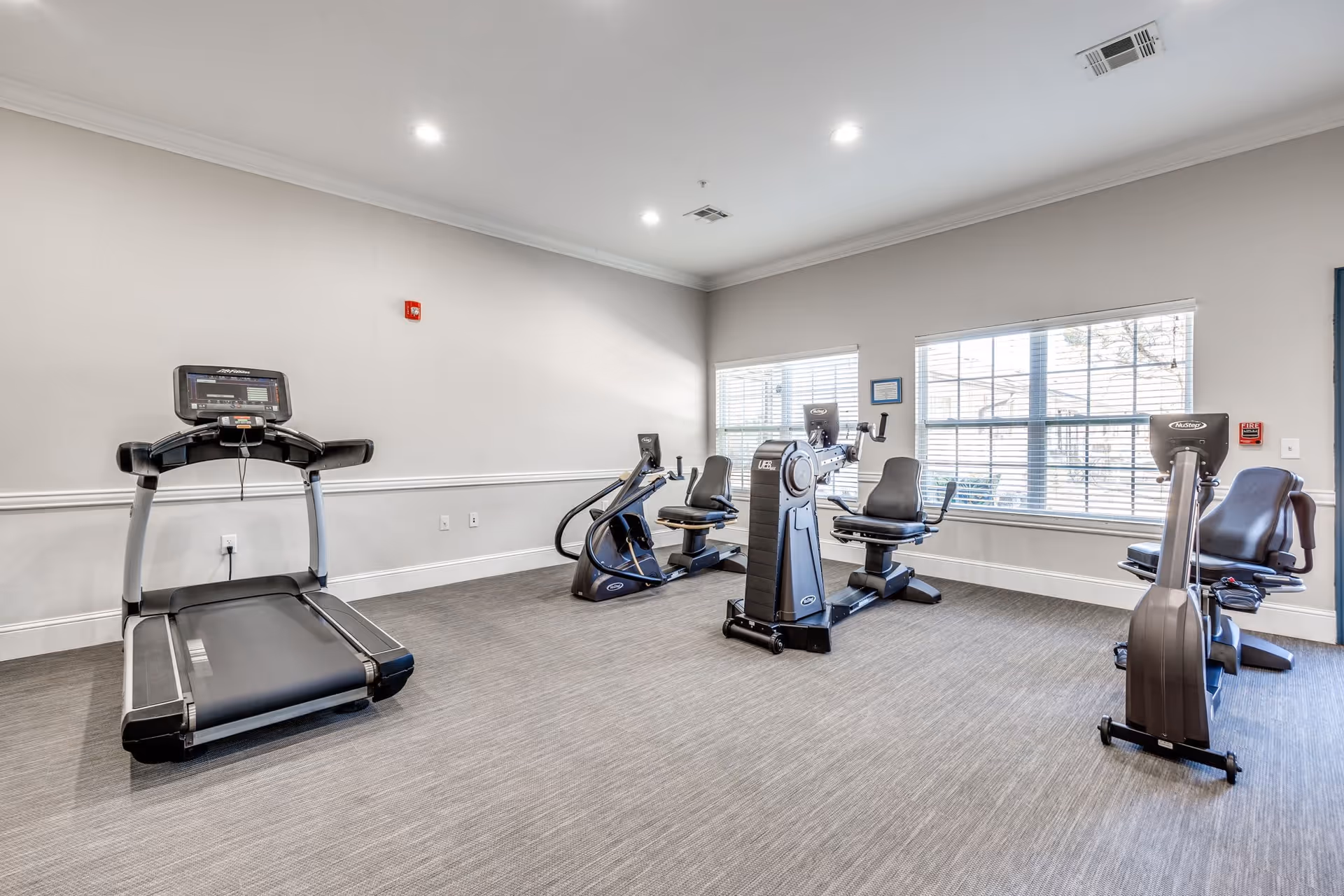 A bright exercise room with a treadmill, two recumbent stationary bikes, and an elliptical machine. The room has large windows with white blinds, light gray walls, and recessed ceiling lights. The floor is covered with gray carpet.