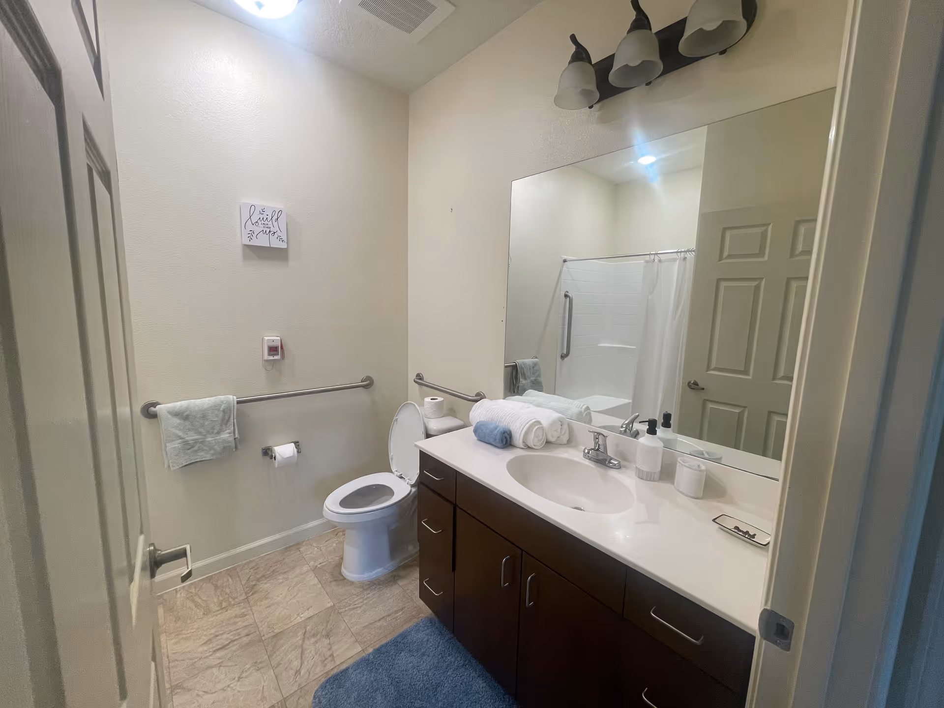 Accessible bathroom with a toilet, vanity and sink, large mirror, grab bars, shower with curtain, and folded towels.