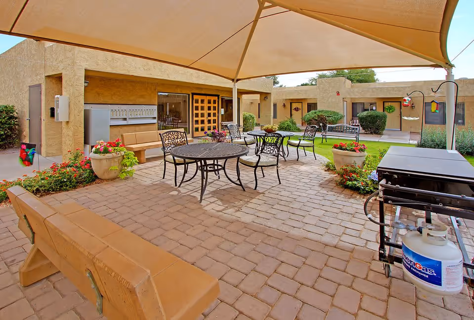 Outdoor patio area at Tempe Post Acute with a large canopy providing shade over metal tables and chairs. There are wooden benches, potted plants with flowers, a propane grill, and a well-maintained garden with bushes and grass surrounding the paved patio. The building exterior is visible in the background with doors and windows.