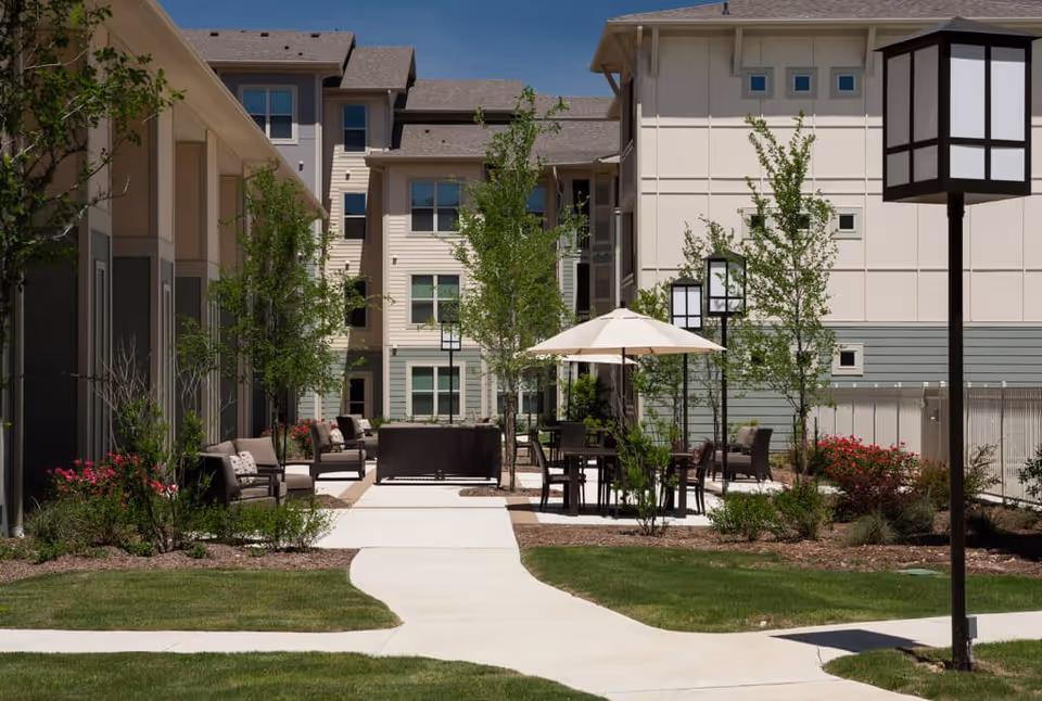 Outdoor courtyard area at Discovery Village Stone Oak with paved walkways, patio tables with umbrellas, chairs, benches, young trees, and landscaped greenery surrounded by multi-story residential buildings.