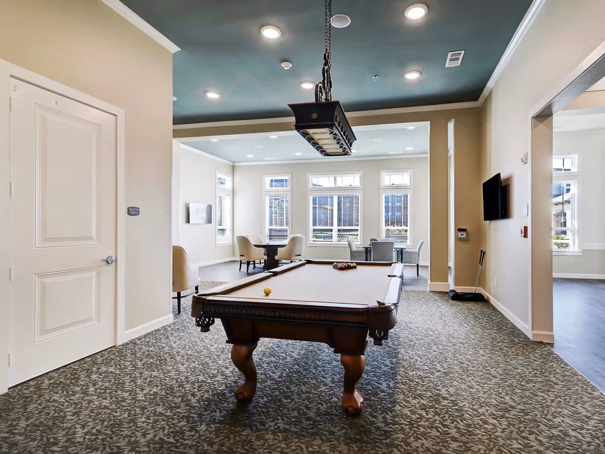 A well-lit recreational room with a pool table in the center, a hanging light fixture above it, and several chairs and tables near large windows letting in natural light. The room has a carpeted floor and neutral-colored walls with a teal ceiling.