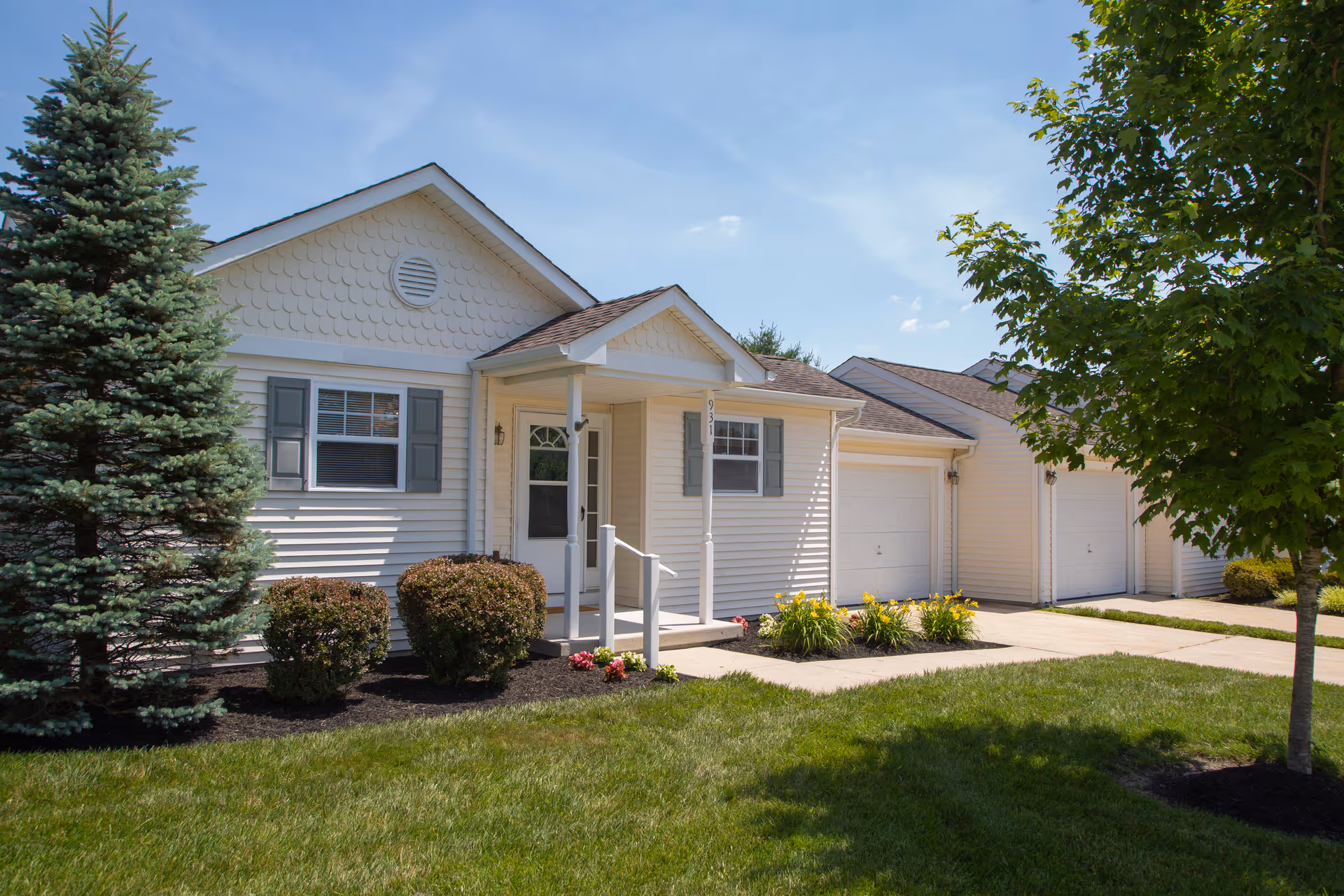 Front exterior of a single-story light-colored residential unit with a small covered porch, attached garages, and landscaped lawn.