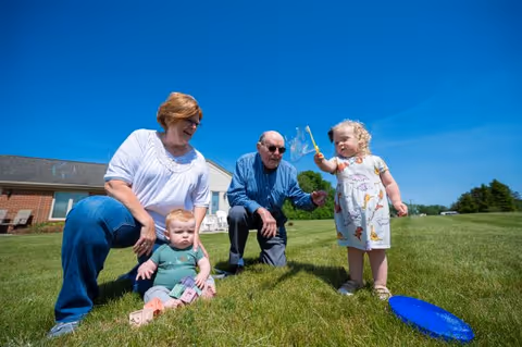 An elderly woman and man outdoors on a sunny day with two young children playing on the grass. The woman is kneeling next to a baby sitting on the grass with toy blocks, while the man is crouching and interacting with a toddler holding a bubble wand. A blue frisbee lies on the grass nearby, and a building is visible in the background under a clear blue sky.