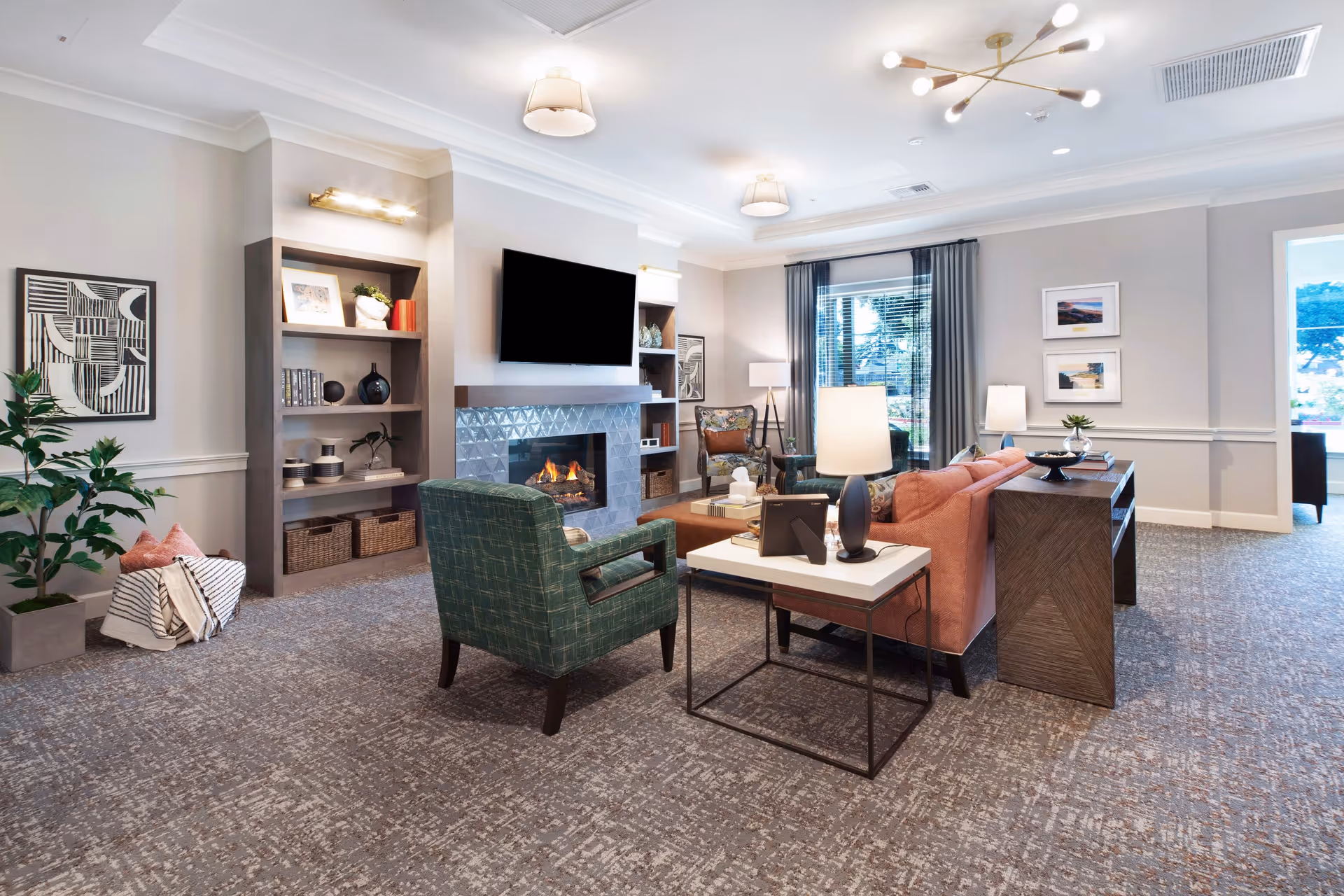 A cozy living room with a fireplace, a wall-mounted TV above it, and built-in shelves on either side. The room features a green armchair, a pink sofa, a side table with a lamp, and large windows with curtains letting in natural light. The carpeted floor and modern light fixtures add to the comfortable ambiance.