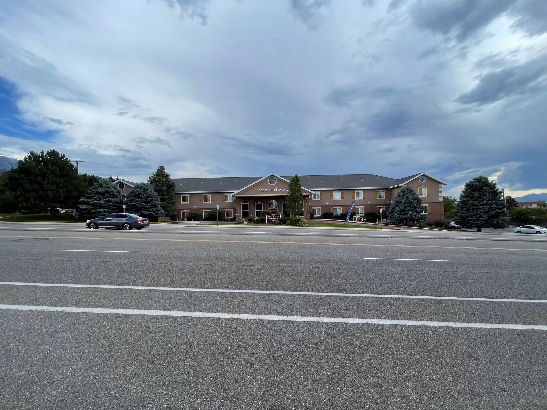 Exterior front view of a two-story assisted living facility named Alta Ridge Assisted Living of Sandy, with a wide road in the foreground, several cars parked along the street, and trees surrounding the building under a cloudy sky.