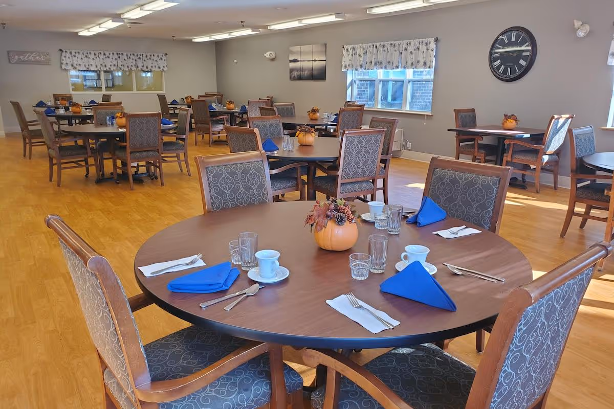 A dining room with multiple round tables set with blue napkins, white cups and saucers, glasses, and silverware. Each table has a small pumpkin centerpiece decorated with autumn leaves and berries. The room has wooden flooring, gray walls, windows with floral valances, and a large clock on one wall.