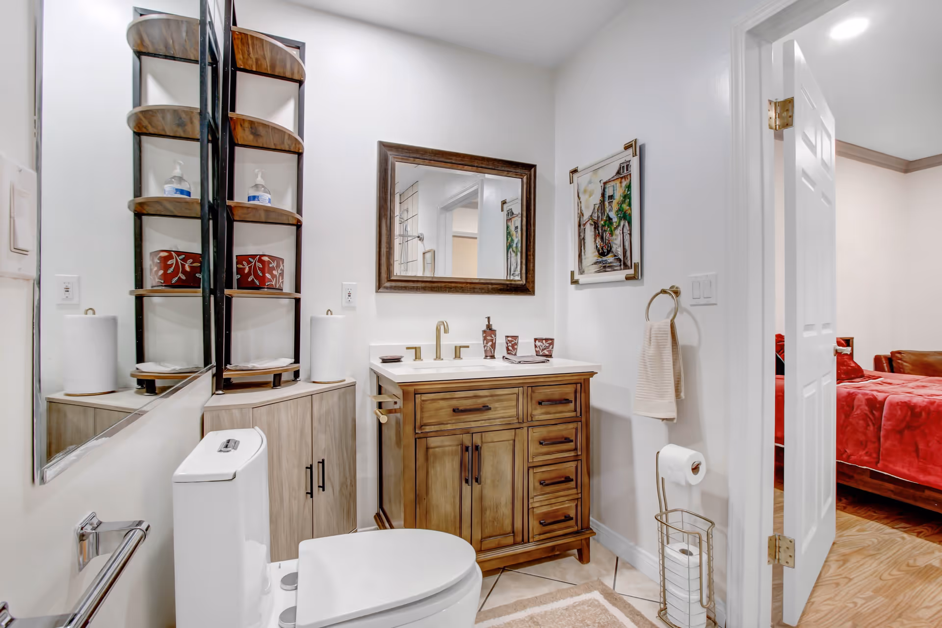 A clean and well-organized bathroom with a white toilet, wooden vanity with a white countertop and gold faucet, a large framed mirror above the sink, a corner shelf holding hand soap and decorative items, a towel ring with a beige hand towel, and a toilet paper holder with extra rolls. The bathroom door is open, showing a glimpse of a bedroom with a red bedspread.