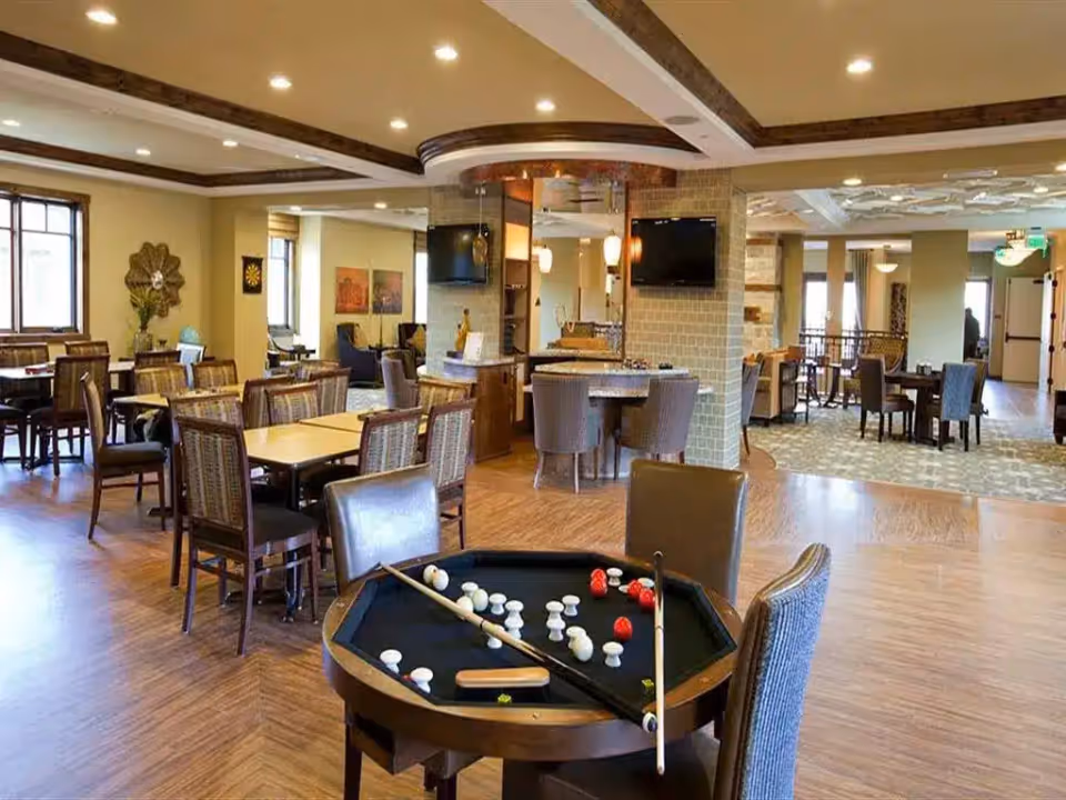 A spacious and well-lit common area in a senior living facility featuring multiple tables and chairs arranged for dining or social activities. In the foreground, there is a round game table with billiard balls and cues. The room has wooden flooring, beige walls, and a ceiling with recessed lighting and decorative beams. There are two wall-mounted televisions and various seating areas visible in the background.
