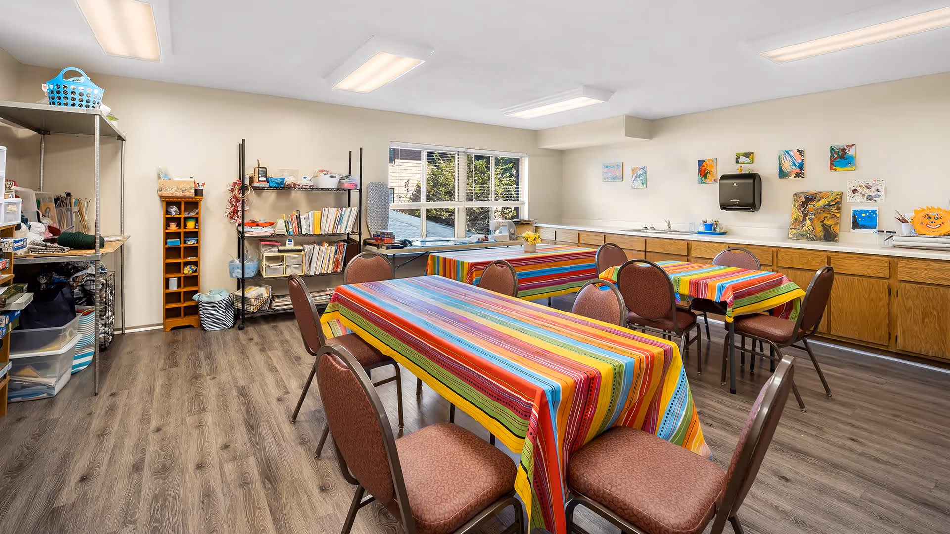 A bright activity room with several tables covered in colorful striped tablecloths and surrounded by chairs. The room has wooden flooring, shelves with books and craft supplies, a large window letting in natural light, and artwork displayed on the walls above a long counter with cabinets and a paper towel dispenser.