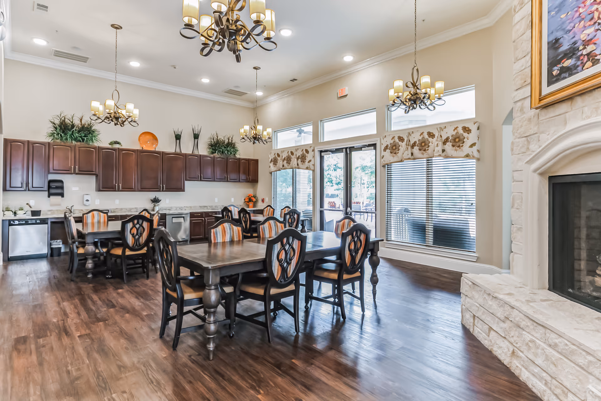 A bright and spacious dining room with wooden floors, multiple dark wood tables and chairs with patterned cushions, large windows with floral valances, and a stone fireplace on the right. The back wall features dark wood cabinets, a dishwasher, and a sink area with decorative plants and vases on top of the cabinets. Elegant chandeliers hang from the ceiling.