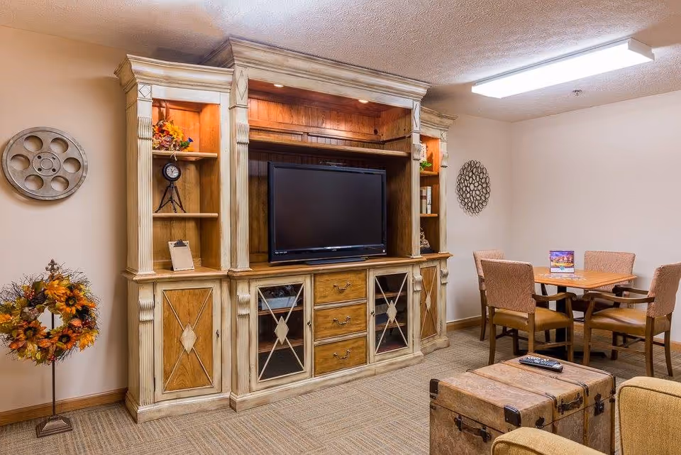 Cozy communal living room with a large wooden entertainment center and TV, a small dining table with chairs, and decorative accents.
