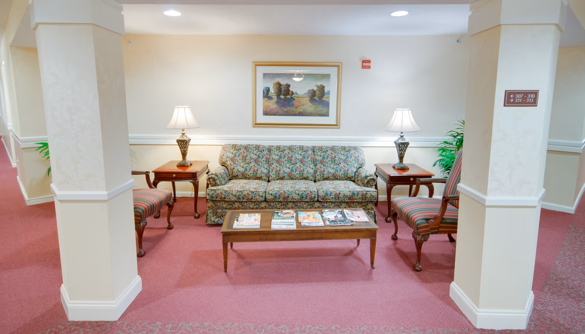A cozy seating area in a senior living facility hallway featuring a floral-patterned sofa, two striped armchairs, two side tables with lamps, a wooden coffee table with magazines, and a framed landscape painting on the wall. The floor is carpeted in pink, and there are two columns framing the seating area.