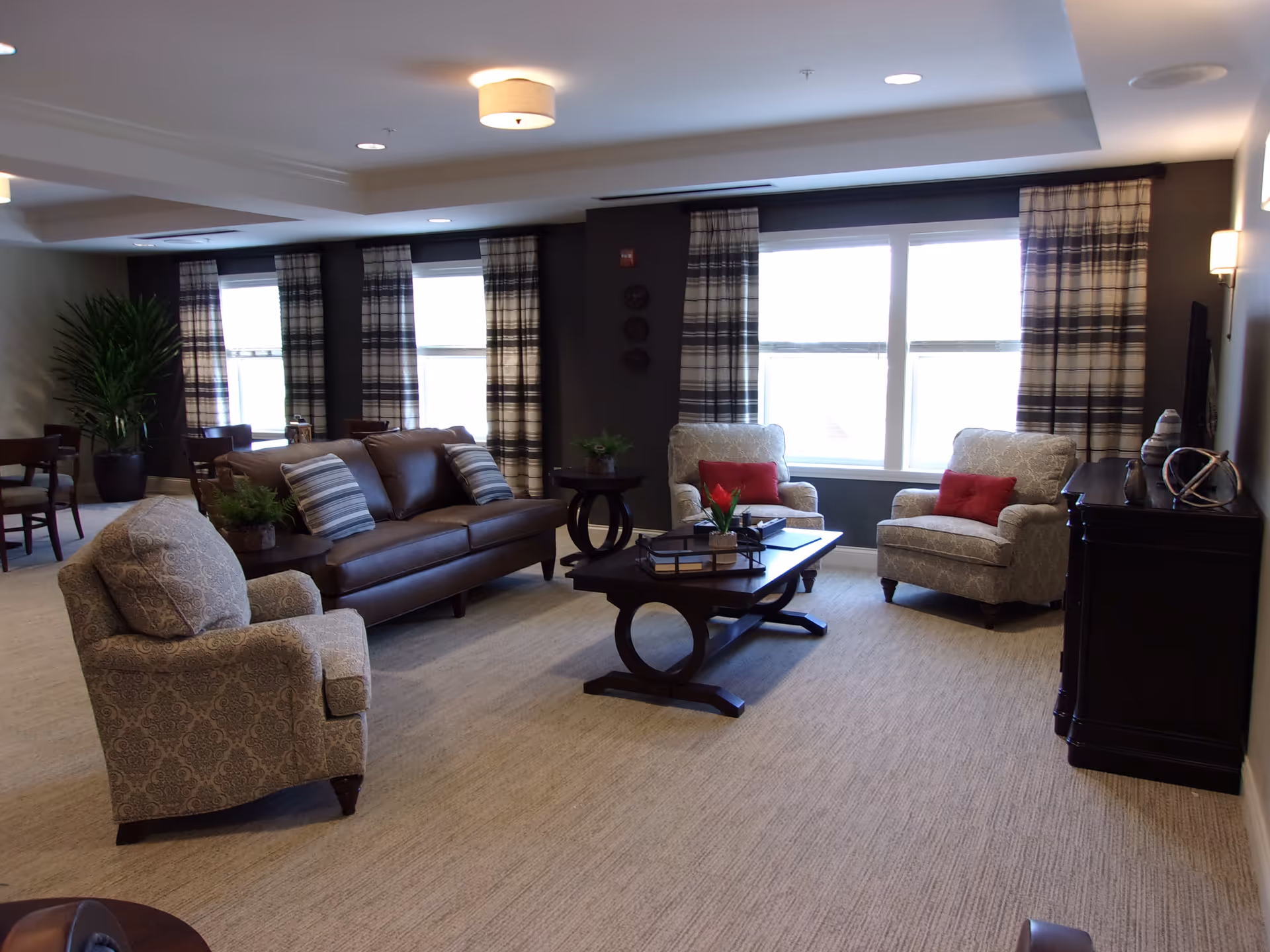A cozy living room area with a brown leather sofa, two patterned armchairs with red cushions, a dark wooden coffee table with decorative items, and large windows with plaid curtains letting in natural light. The room has beige carpeting and neutral-colored walls with ceiling lights.