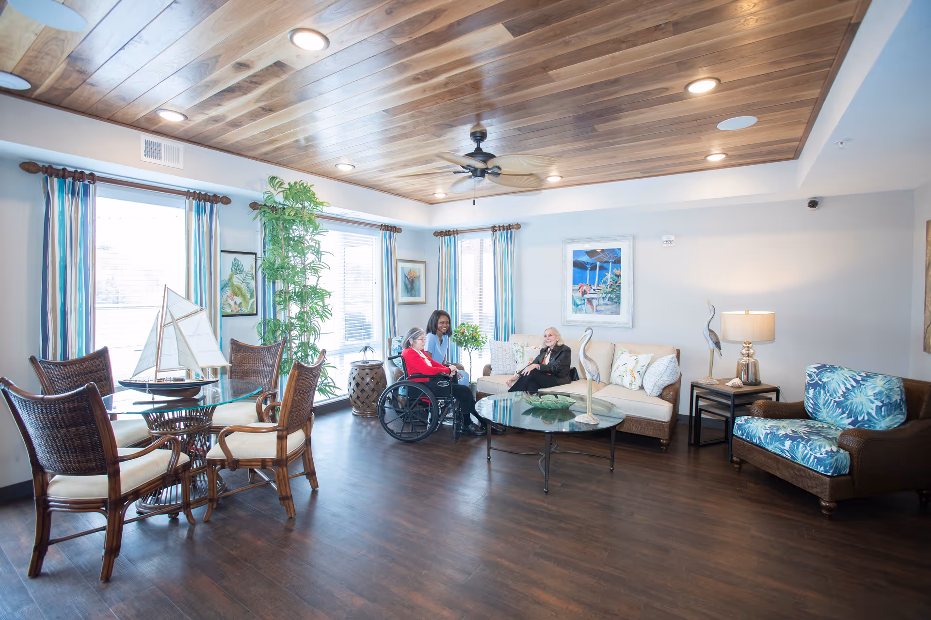 A bright and spacious senior living common area with wooden flooring and a wooden ceiling featuring recessed lighting and a ceiling fan. The room has large windows with striped curtains, a round glass dining table with four wicker chairs, a beige sofa with decorative pillows, a brown armchair with a blue floral cushion, and a glass coffee table with decorative items. Two elderly women, one in a wheelchair, and a caregiver are seated and engaged in conversation.