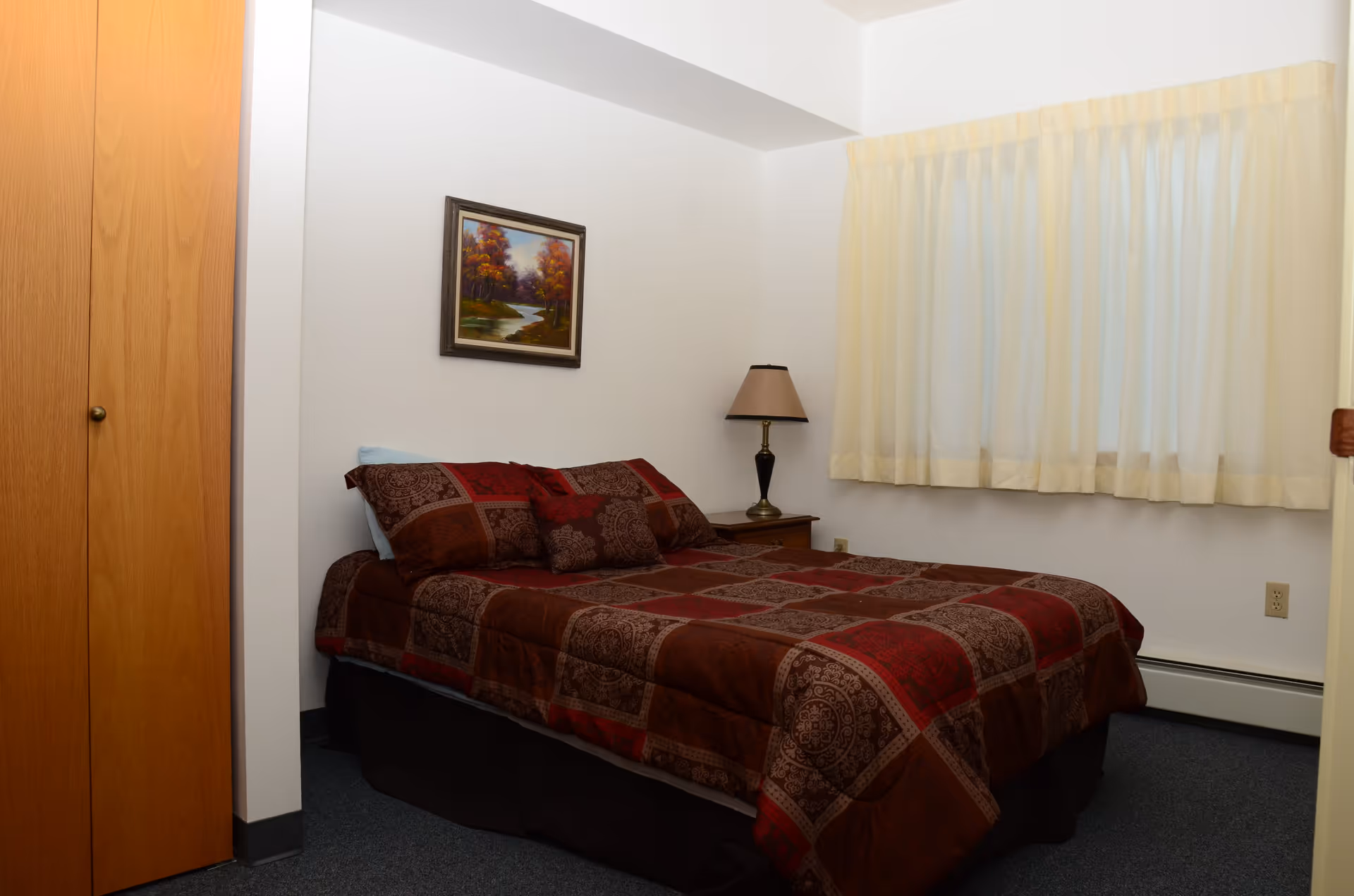A simple bedroom with a bed covered in a red and brown patterned comforter, a wooden nightstand with a lamp, a framed painting of a river and trees on the wall, a window with cream-colored curtains, and a wooden closet door.