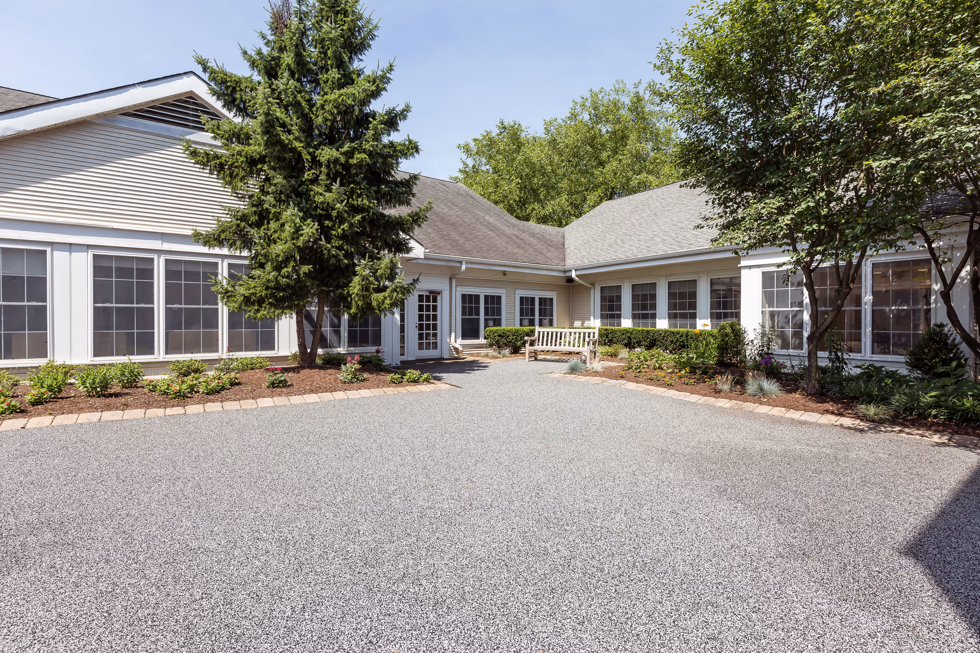 Outdoor courtyard area of a senior living facility with a paved ground, landscaped garden beds, trees, shrubs, and a wooden bench near the building entrance. The building has large windows and light-colored siding under a gray roof.