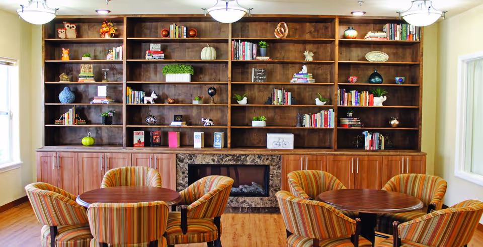 Cozy common room with large built-in wooden bookshelves, a fireplace, and round tables surrounded by striped upholstered chairs.