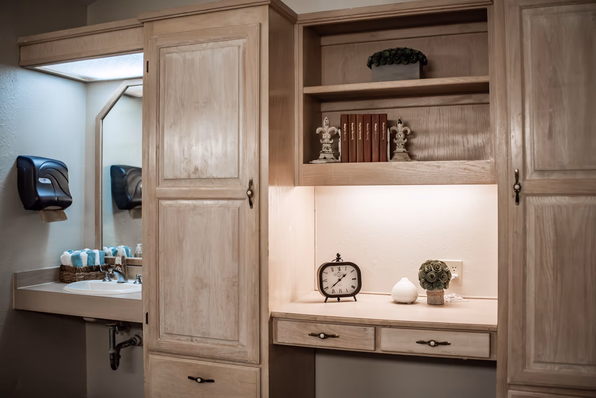 A bathroom sink area with a large mirror, a paper towel dispenser on the wall, and a basket of rolled towels. Adjacent to the sink is a built-in wooden cabinet with shelves holding decorative items including books, a clock, and small ornaments.