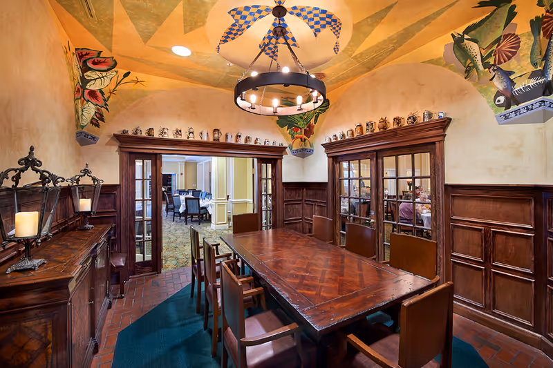 A formal dining room with a long wooden table surrounded by eight wooden chairs. The room features wood-paneled walls, decorative ceiling paintings with colorful designs, and a circular chandelier with candle-style lights hanging from the ceiling. There are two sets of glass-paneled double doors leading to another dining area with tables and chairs.