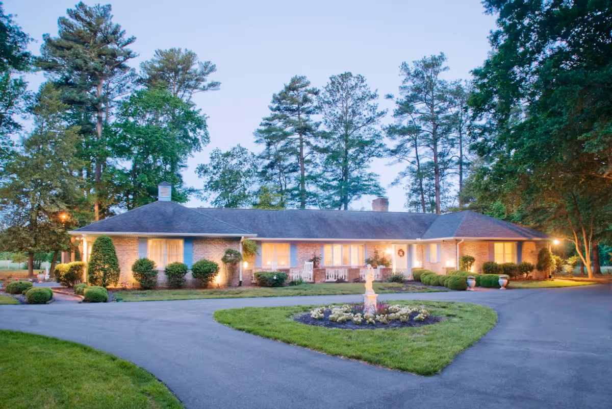 Exterior view of a single-story brick building surrounded by trees and well-maintained landscaping, with a circular driveway and a flower bed with a small statue in the center, taken during dusk with lights illuminating the building.