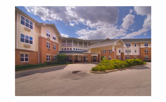 Front entrance of a multi-story senior living building with a covered porte-cochere, landscaped island, and cloudy sky.