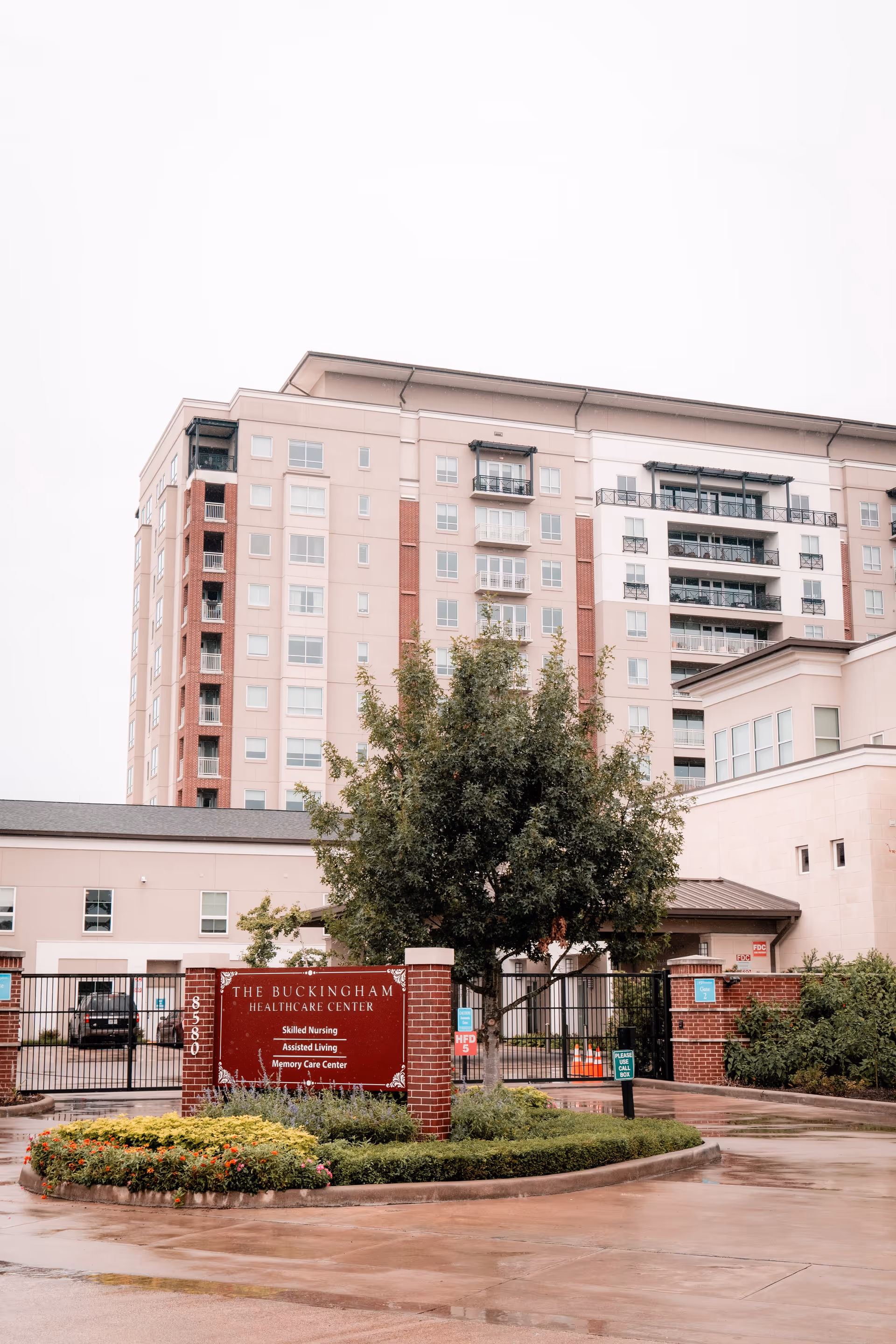 Exterior view of The Buckingham Healthcare Center, a multi-story building with balconies and windows. In front, there is a red sign with white text indicating services such as Skilled Nursing, Assisted Living, and Memory Care Center. The area around the sign has neatly trimmed bushes and flowers, and the ground appears wet from recent rain.