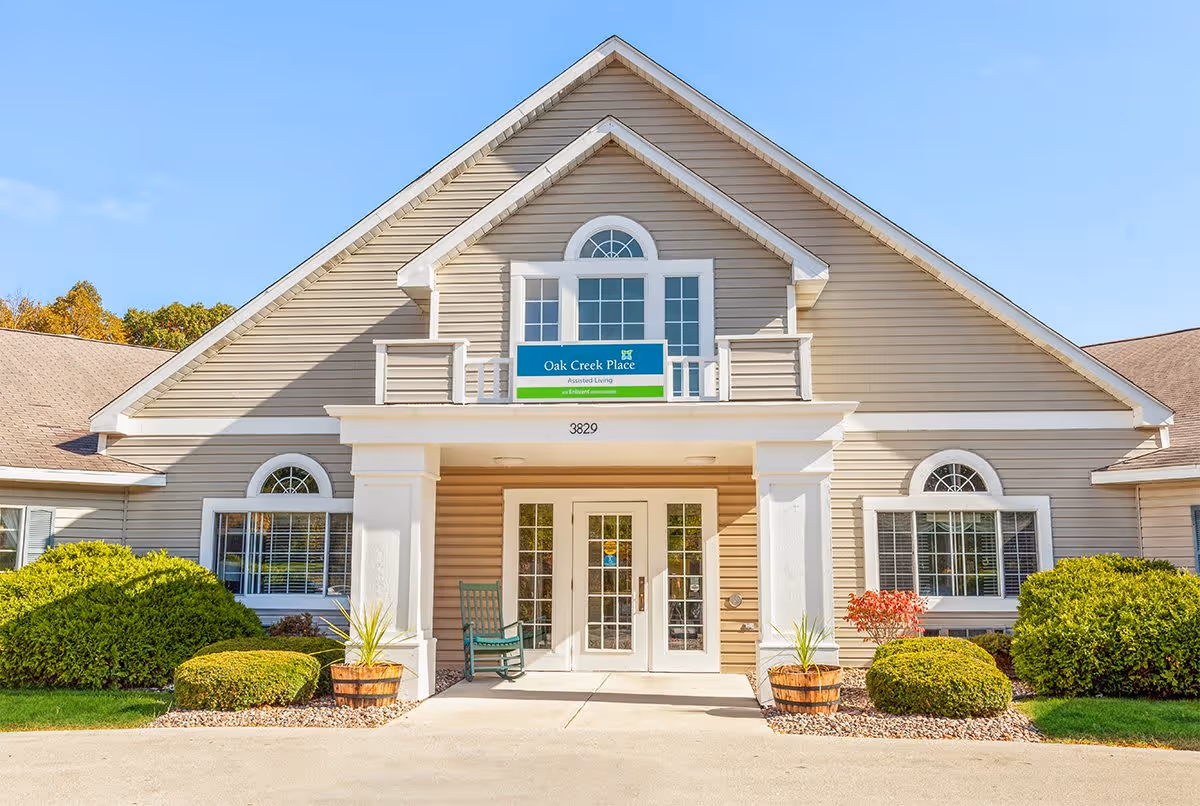 Front exterior view of a beige building with white trim and a peaked roof, featuring a sign that reads 'Oak Creek Place Assisted Living'. There are two large windows with white frames on either side of the entrance, which has double glass doors and a small porch with two white columns. Green shrubs and plants are neatly arranged around the entrance.
