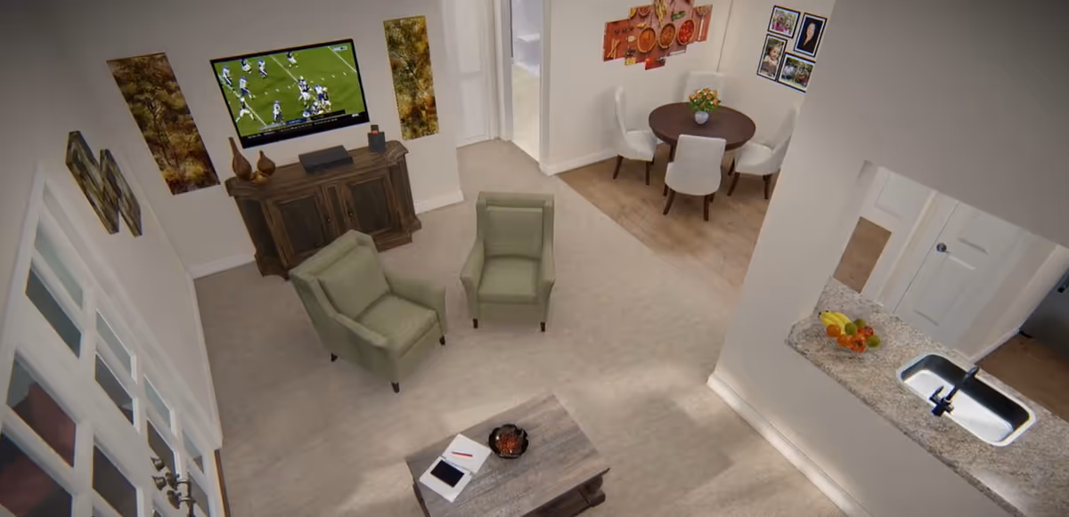 Open living area with two green armchairs facing a wall-mounted TV, a coffee table, a nearby round dining table, and a kitchen counter with sink and fruit bowl.