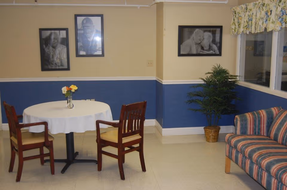 Small communal dining area with a round table and chairs, framed photos on the wall, a potted plant, and a striped sofa.