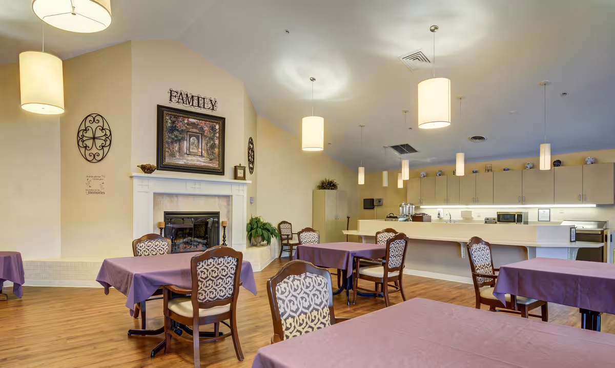A dining area in Belmont Healthcare Center with several tables covered in purple tablecloths and wooden chairs with patterned cushions. The room features a fireplace with a framed picture above it and the word 'FAMILY' displayed on the wall. Pendant lights hang from the ceiling, and there is a kitchen counter with cabinets and appliances in the background.