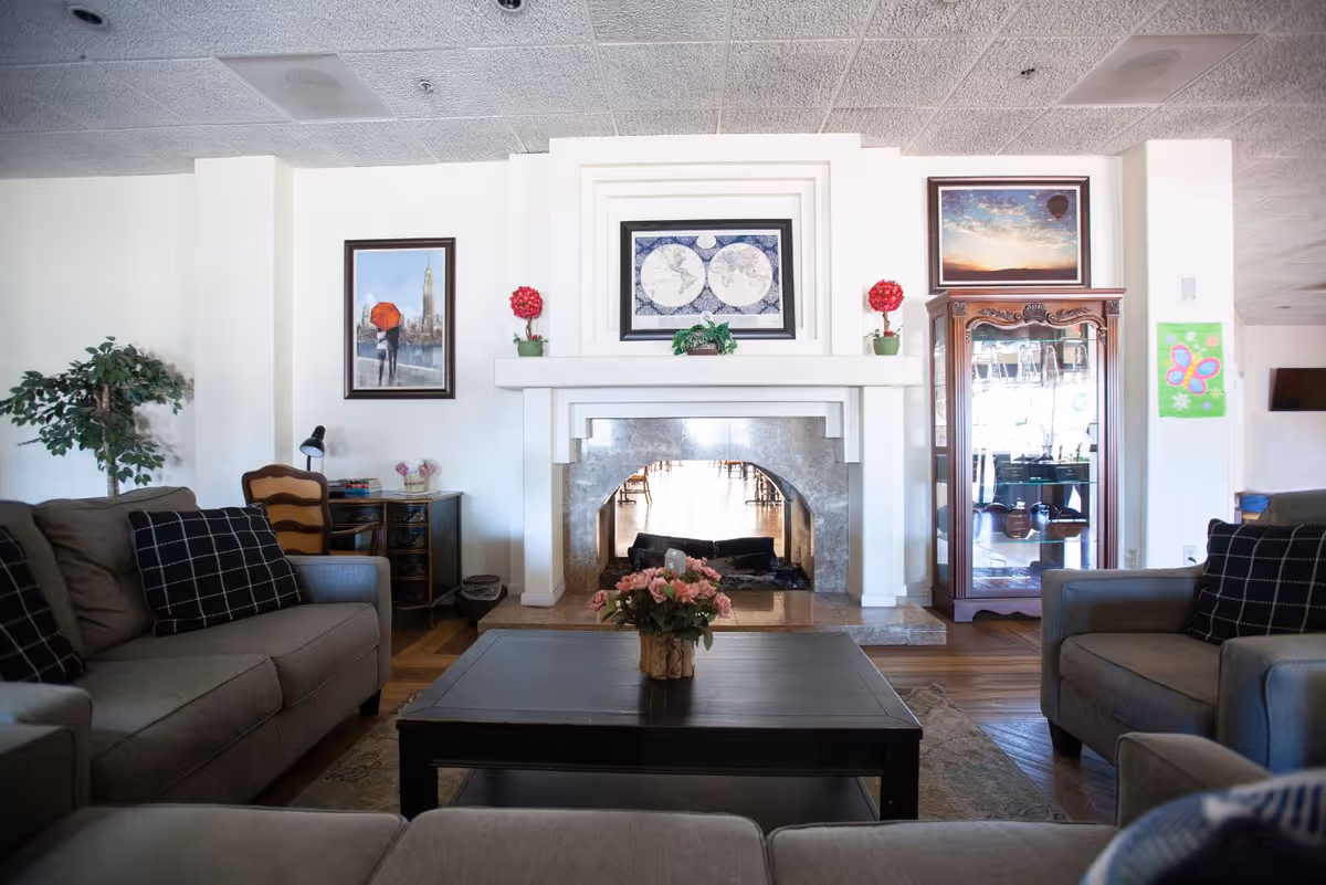 A cozy living room with gray sofas and black checkered pillows arranged around a dark wooden coffee table with a flower arrangement. The room features a white fireplace with a framed world map above it, flanked by two red floral decorations. There is a glass display cabinet on the right and framed artwork on the walls. A potted plant and a small desk with a chair are visible on the left side.