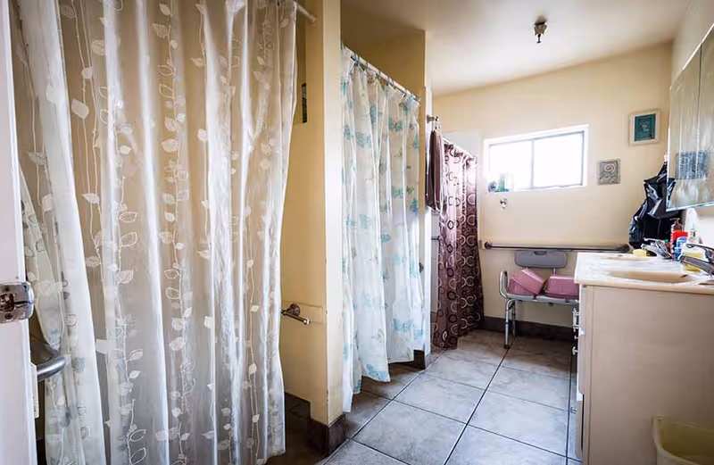 A bathroom with multiple shower stalls separated by curtains, a window letting in natural light, a sink with a countertop, and a shower chair with pink cushions.