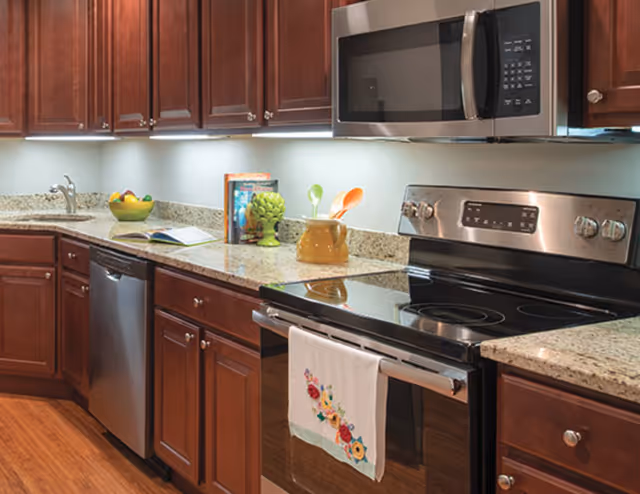 Modern kitchen with wooden cabinets, granite countertops, stainless steel appliances including a microwave, stove, and dishwasher. A colorful towel hangs on the oven handle, and various kitchen items such as a bowl of fruit, cookbooks, and utensils are placed on the counter.