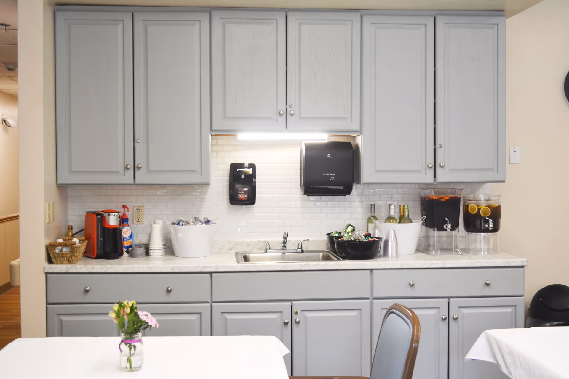 A kitchenette area with light gray cabinets and a white marble countertop. On the countertop, there is a coffee maker, disposable cups, a basket with condiments, a sink, a bowl with canned drinks, bottles of wine in an ice bucket, and two large beverage dispensers with drinks and lemon slices. In front of the kitchenette, there is a table with a small vase of flowers and a chair.