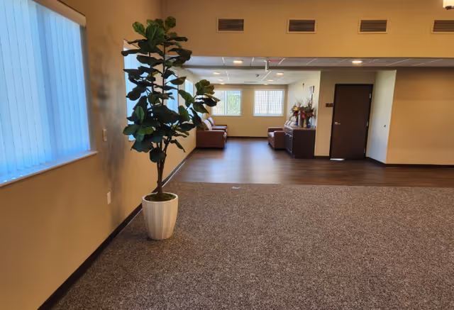 Carpeted common area inside a memory care facility with a potted plant in the foreground and a seating area and door in the background.