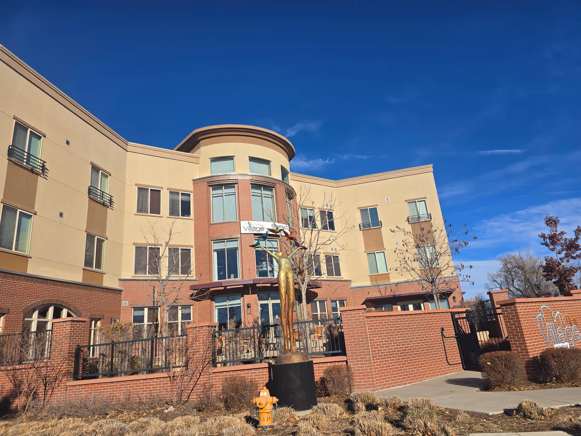 Exterior view of a multi-story senior living facility building with beige and brick facade under a clear blue sky. In front of the building is a bronze statue of a person with arms raised, surrounded by a brick fence and some leafless trees and bushes. A yellow fire hydrant is visible near the sidewalk.