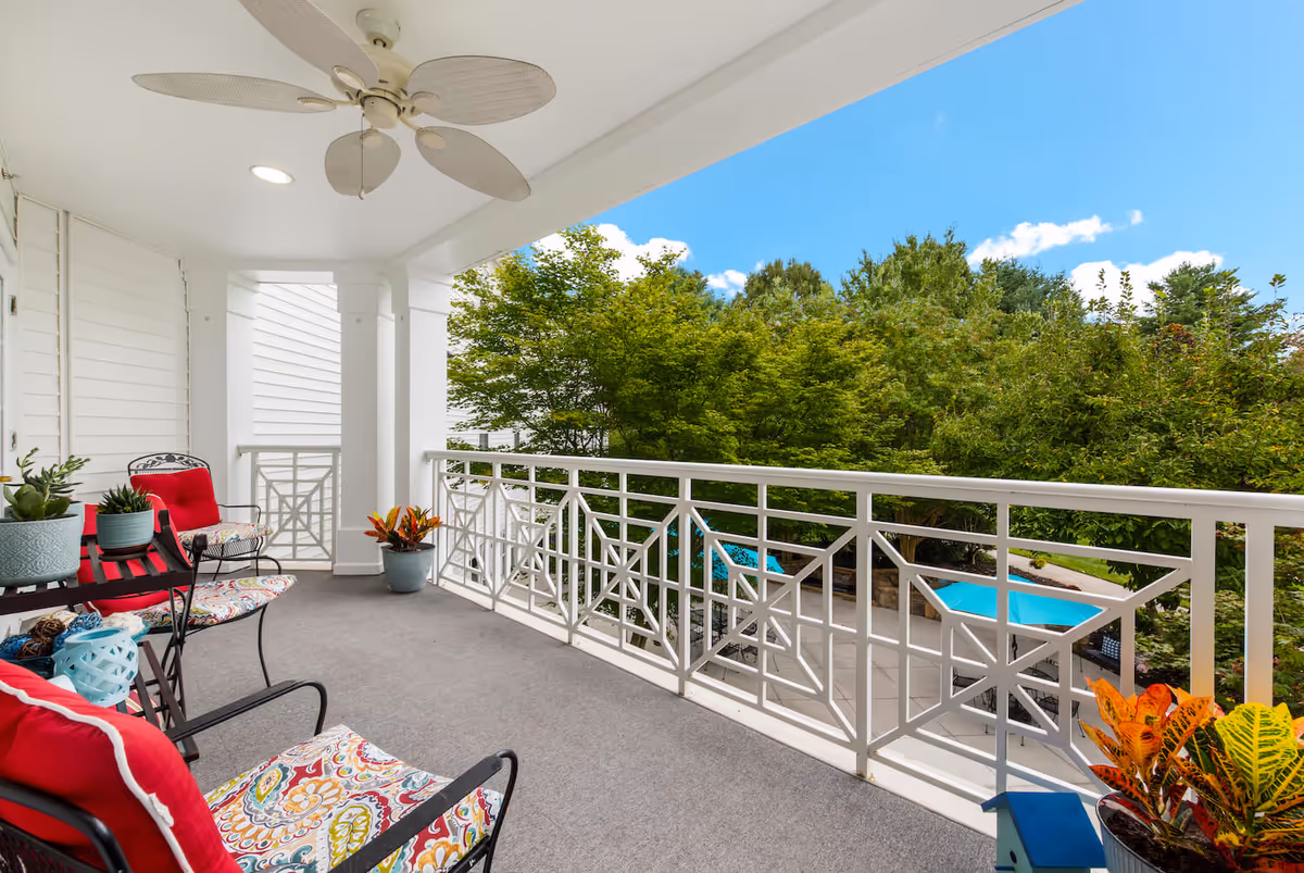 Covered balcony with colorful cushioned chairs, potted plants, a ceiling fan and white railing overlooking trees and a pool area.