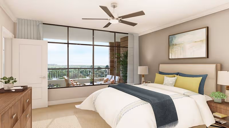 A bright and spacious bedroom with a large bed featuring white bedding, blue and yellow pillows, and a dark blue throw blanket. The room has a wooden dresser with a plant and decor on top, two bedside tables with lamps, a ceiling fan, and a large window with a view of an outdoor patio and greenery beyond.