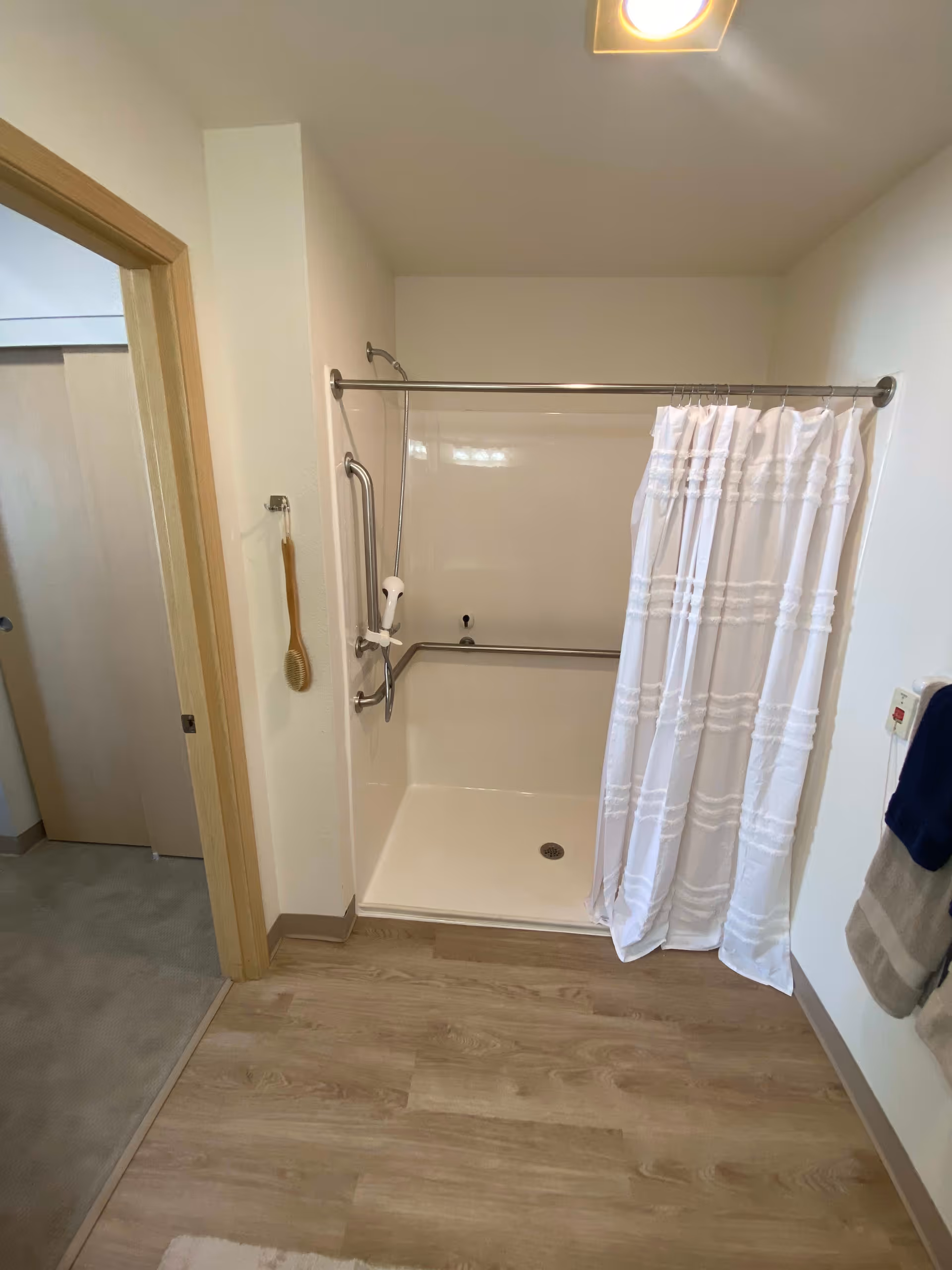 A bathroom shower area with a white shower curtain partially drawn. The shower has grab bars installed on the walls and a handheld showerhead. A wooden bath brush hangs on the wall outside the shower. The floor is wood-patterned, and towels hang on a rack to the right.