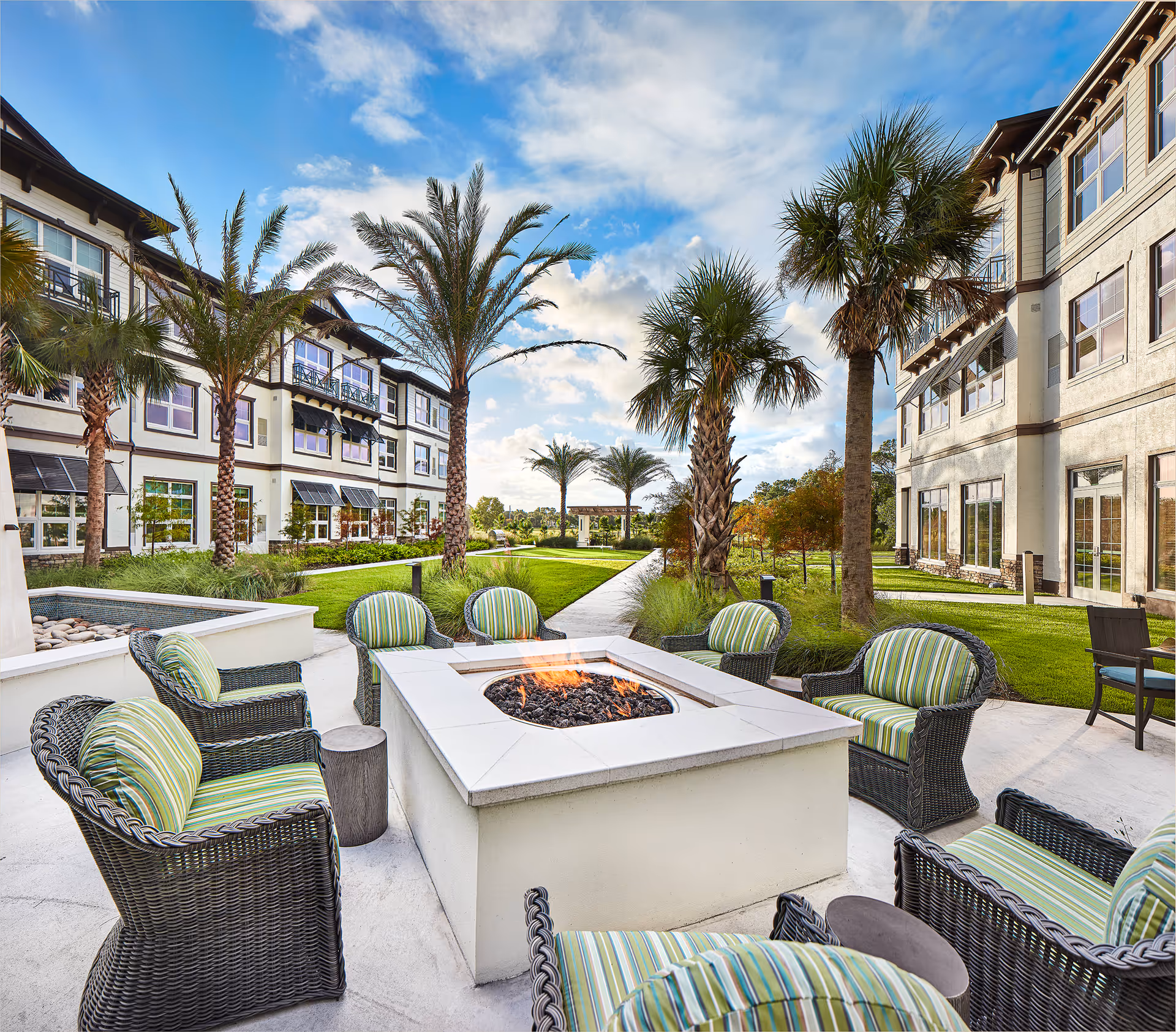 Outdoor seating area with wicker chairs featuring green and white striped cushions arranged around a square fire pit. The area is surrounded by palm trees and well-maintained grass, with two multi-story buildings on either side under a partly cloudy blue sky.