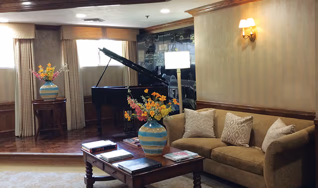 A cozy living room area featuring a brown sofa with patterned pillows, a wooden coffee table with books and a vase of colorful flowers, a grand piano near windows with beige curtains, and a wall lamp providing warm lighting.