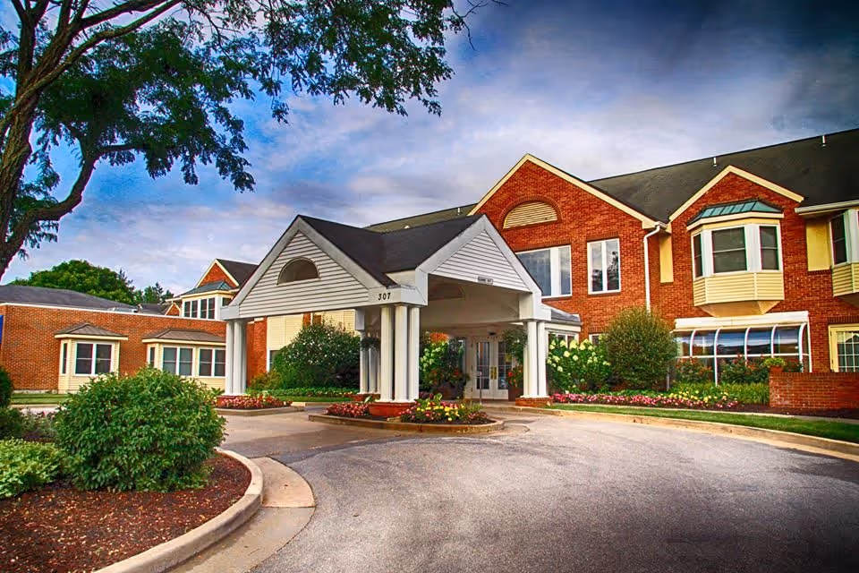 Front exterior view of Brooking Park facility showing a brick building with a covered entrance, landscaped bushes, flowers, and a curved driveway under a partly cloudy sky.