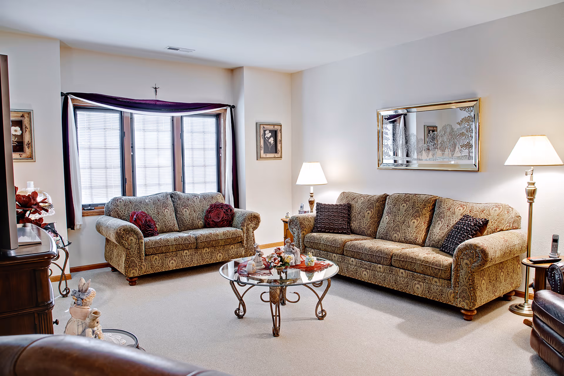 A cozy living room with two patterned sofas facing each other, a glass coffee table with decorative items in the center, a large window with blinds and dark curtains, two floor lamps providing warm light, and framed artwork on the walls.