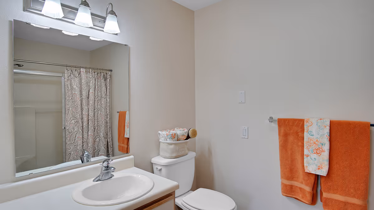Bathroom with sink and mirror, toilet topped with a basket, shower with patterned curtain, and orange towels on a wall rack.