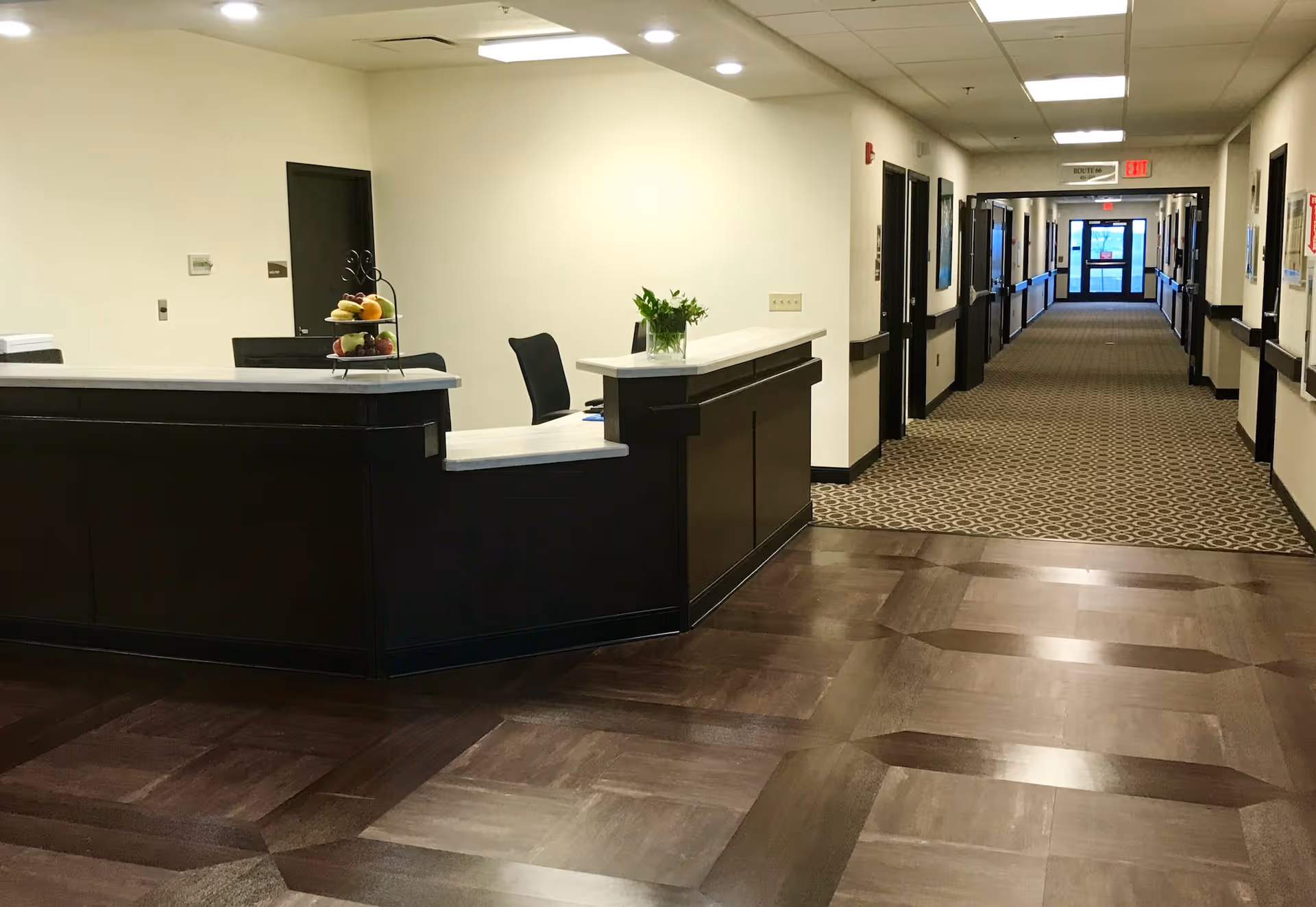 Reception desk area with a black chair, a tiered fruit basket, and a small plant on the counter. The desk is positioned near a long hallway with patterned carpet and multiple doors on each side, leading to an exit door with glass panels at the far end.